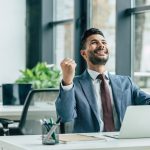 happy-businessman-looking-up-and-showing-winner-gesture-while-sitting-at-workplace.jpg
